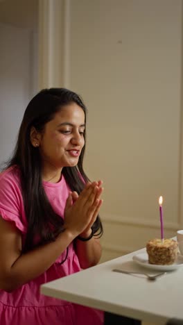 A-happy-brunette-girl-in-a-pink-dress-sits-in-front-of-a-table-in-front-of-a-small-cake-and-blows-out-a-pink-candle-on-it-applauding-and-clapping-her-hands-in-a-modern-apartment